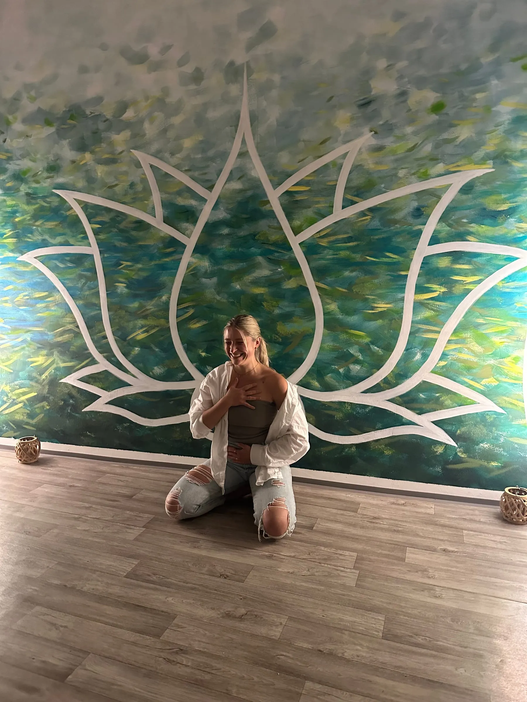 Katerina Kaslova laughing with one hand on her heart, kneeling in front of a painted teal lotus mural
